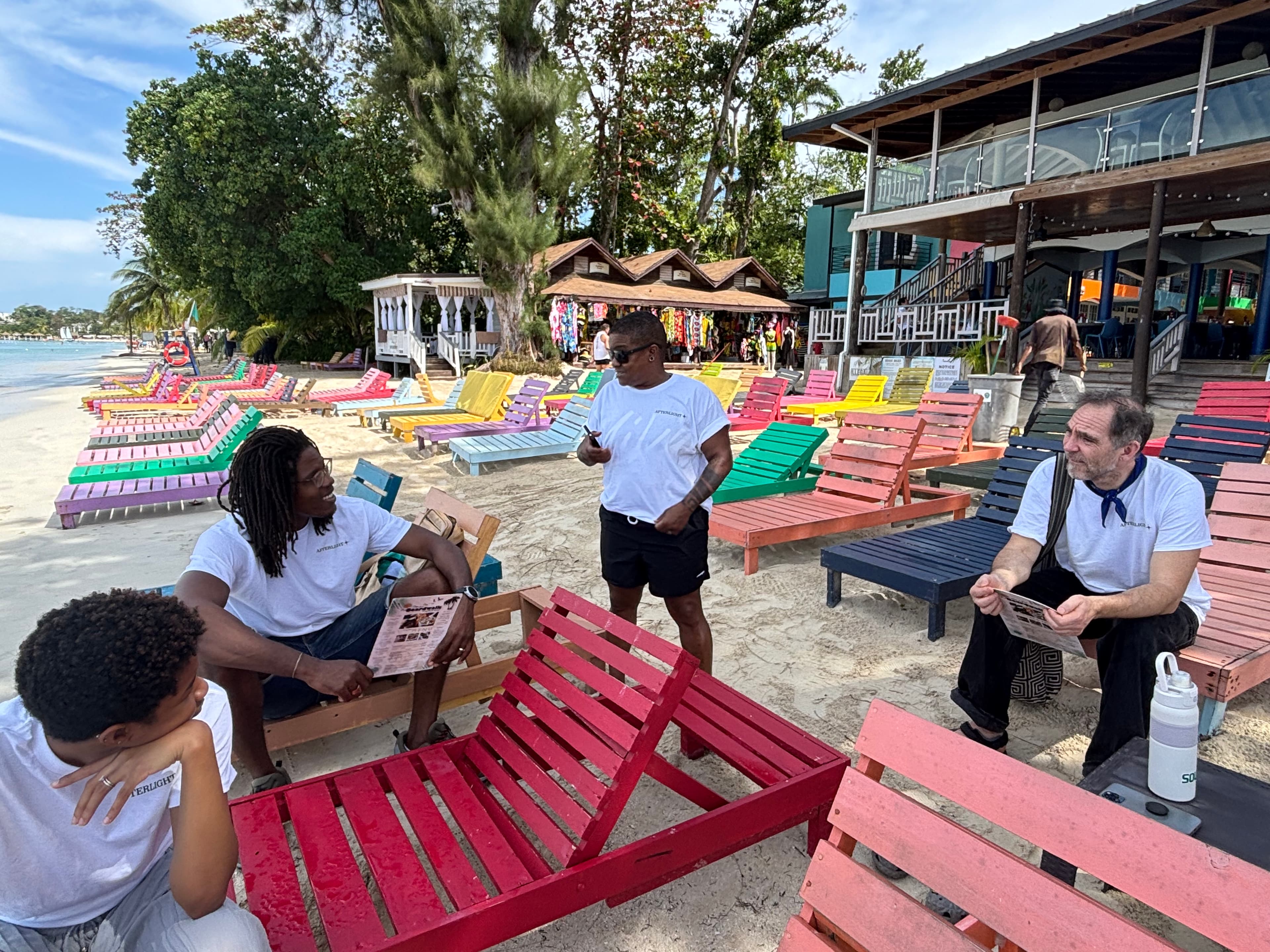 Afterlight team members in relaxed conversation among brightly coloured beach loungers on a Jamaican beachfront, reading and talking together