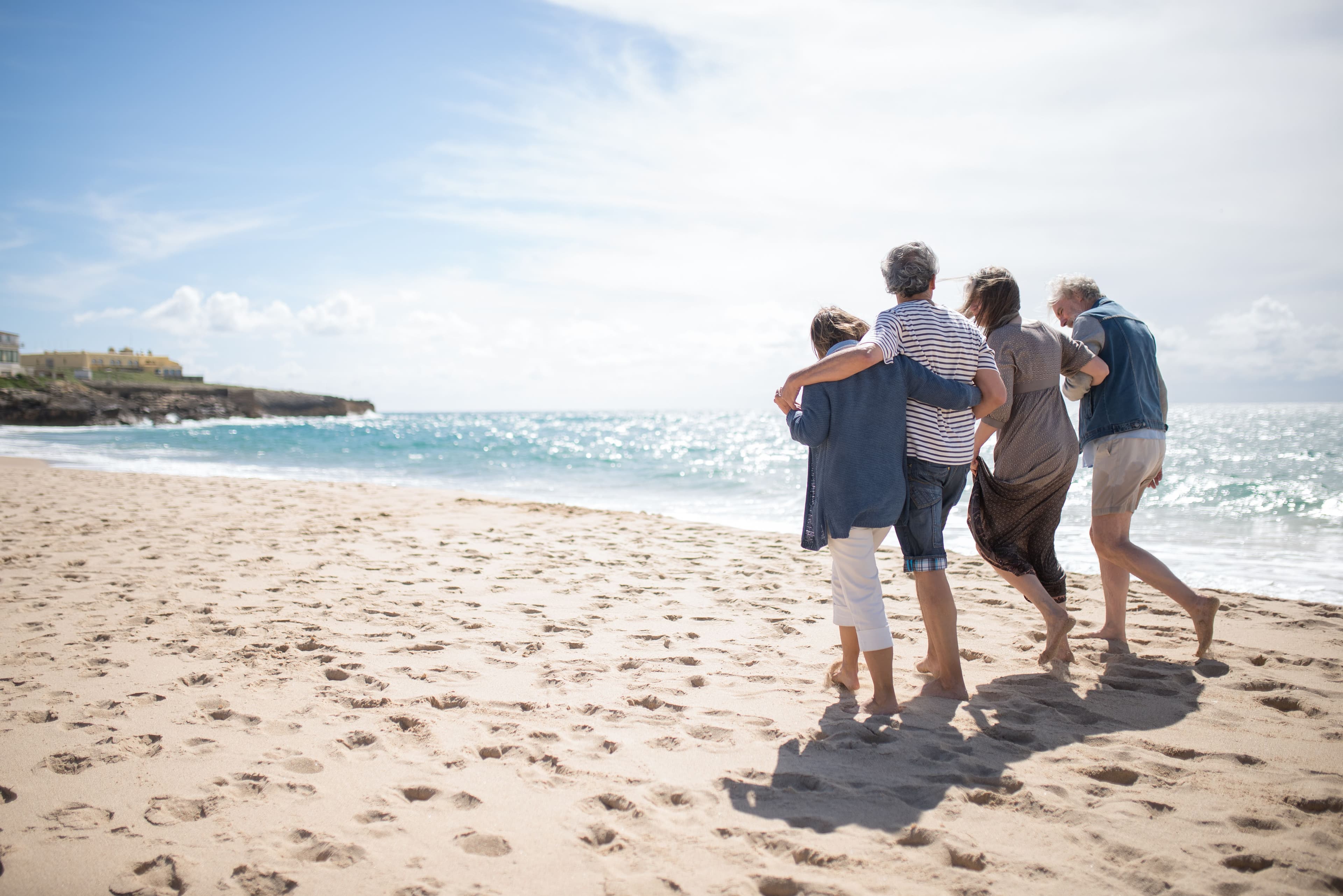 Four people walking arm in arm along a sunlit beach toward the ocean, supporting one another